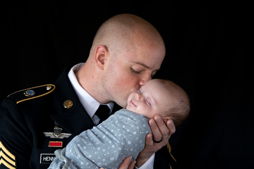 A soldier in dress uniform kisses the head of a swaddled baby.
