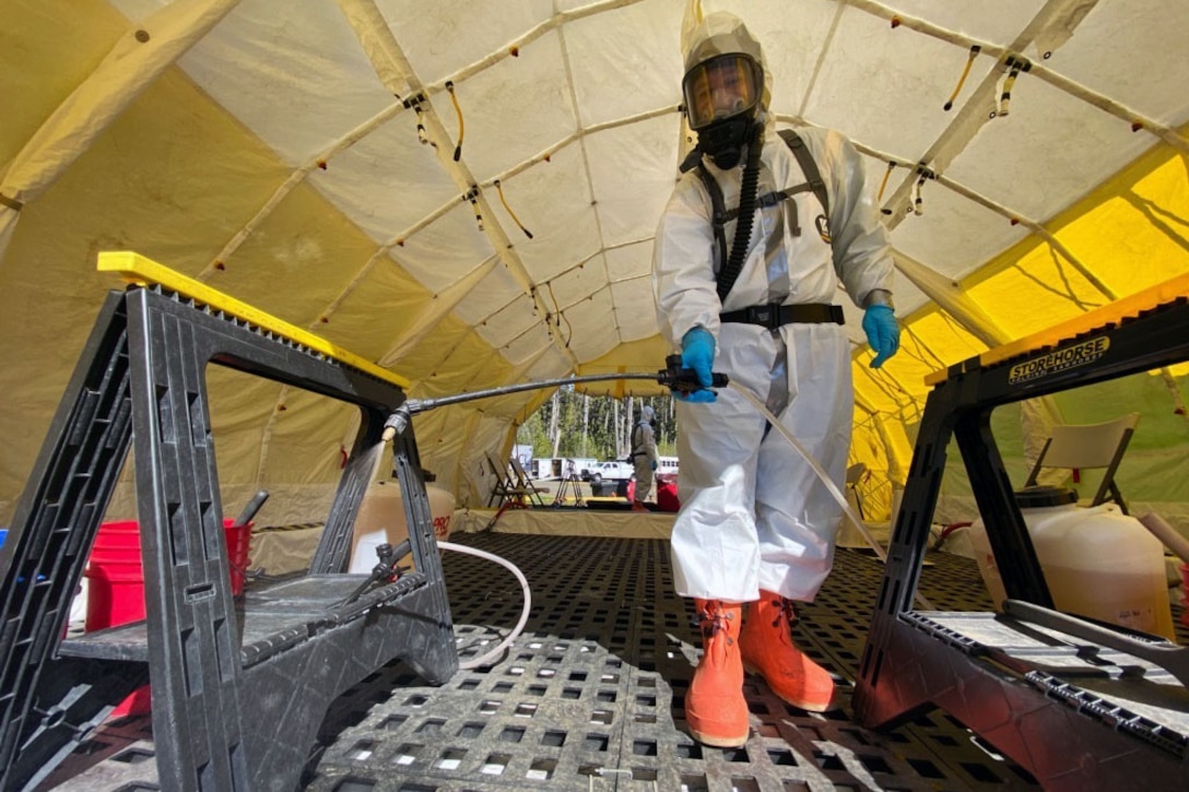 A service member wearing personal protective equipment decontaminates an area inside a tent.