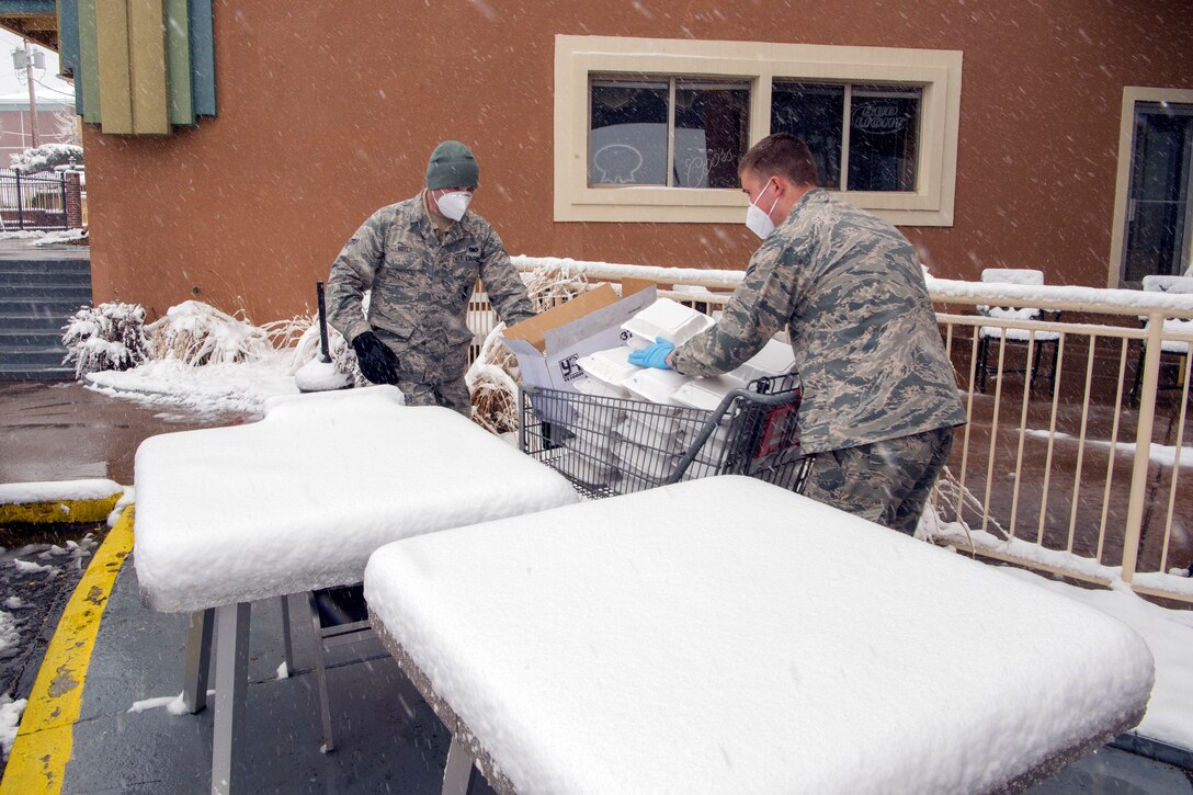 Two service members deliver pre-packaged meals. They are outside a building and there is snow on the ground.