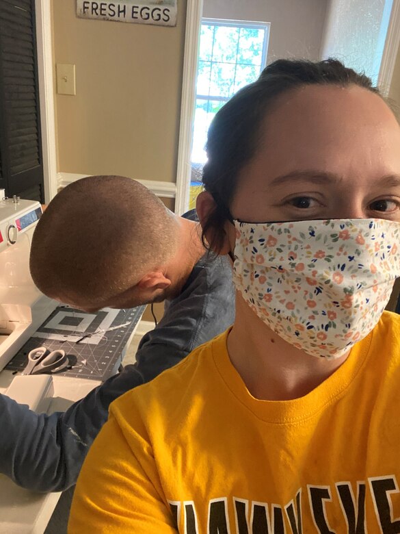 Photo shows woman taking a selfie with her husband in the background sewing a mask.
