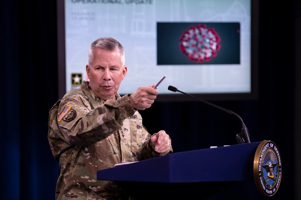 A man in a military uniform stands at a lectern while gesturing with a pen. A graphic of the COVID-19 virus is on a screen in the background.