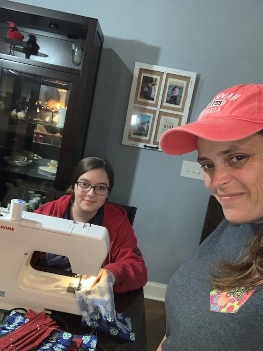 Photo shows young woman sitting at a sewing machine and mother standing next to her taking a photo of both of them.