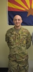 White male in green camouflage uniform stands in front of blue, red and yellow flag with a star in the middle.