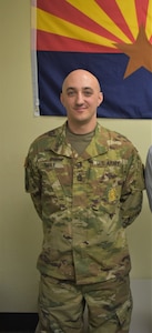 White male in green camouflage uniform stands in front of blue, red and yellow flag with a star in the middle.