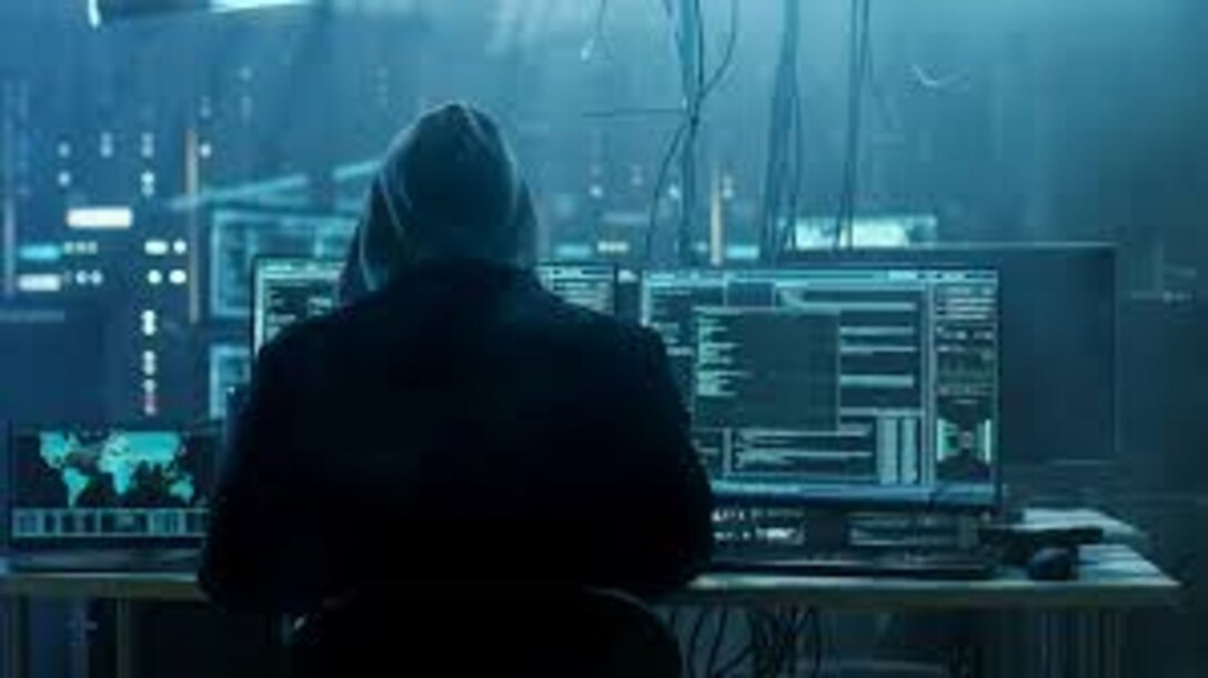 The view from behind a hooded figure, who is looking at several computer screens in a dingy room.