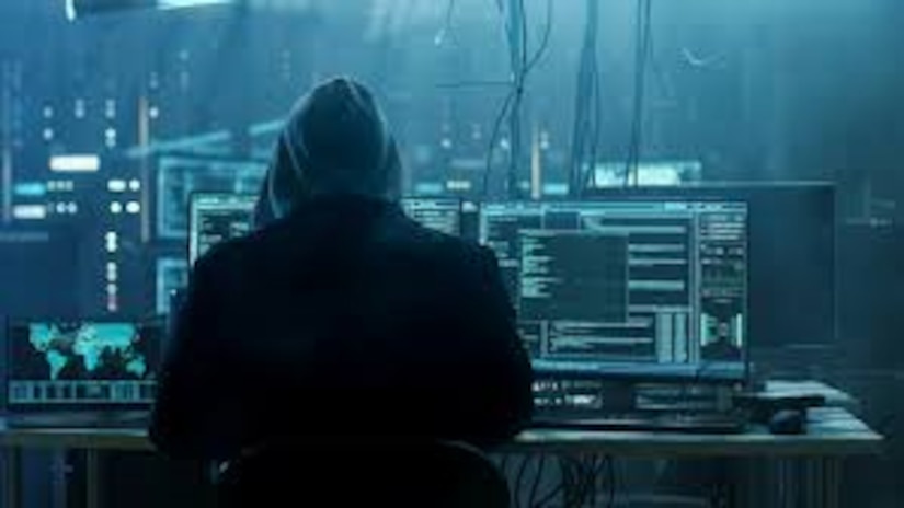The view from behind a hooded figure, who is looking at several computer screens in a dingy room.