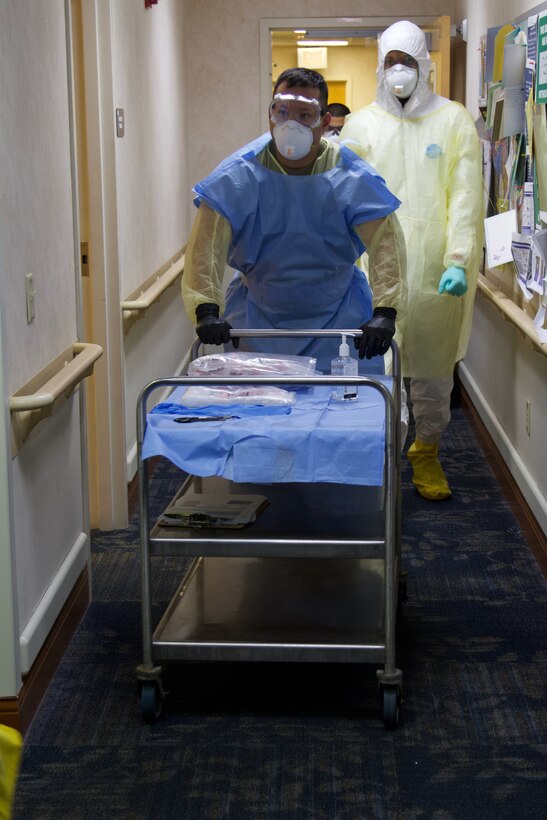 A soldier wearing protective equipment pushes a cart holding COVID-19 testing supplies down a hallway.