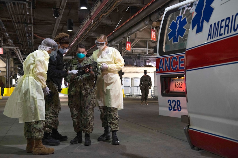 Four sailors dressed in protective gear consult an electronic device as they stand by the open back door of an ambulance.