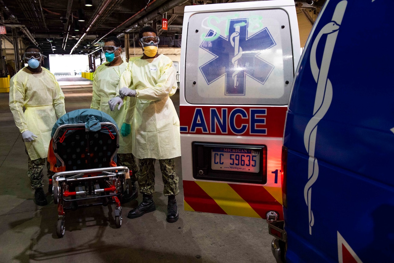 Sailors wearing protective gear stand by the open back door of an ambulance.