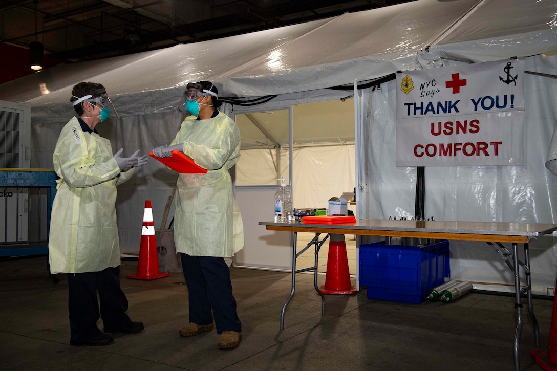 Two Navy officers stand and talk in front of a tent bearing a sign that reads “NYC Thanks You! USNS Comfort.”