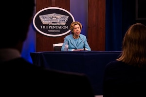 Woman seated at a table with a microphone speaks. There is a sign in the background stating that it is “The Pentagon.”