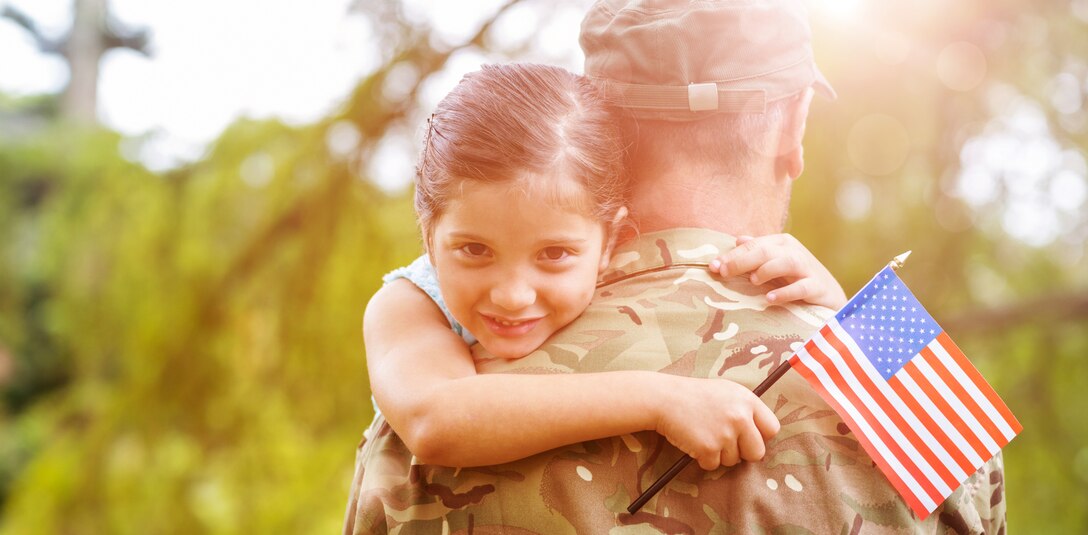 photo shows a young girl hugging her father who is in a military uniform while she is holding an American flag.
