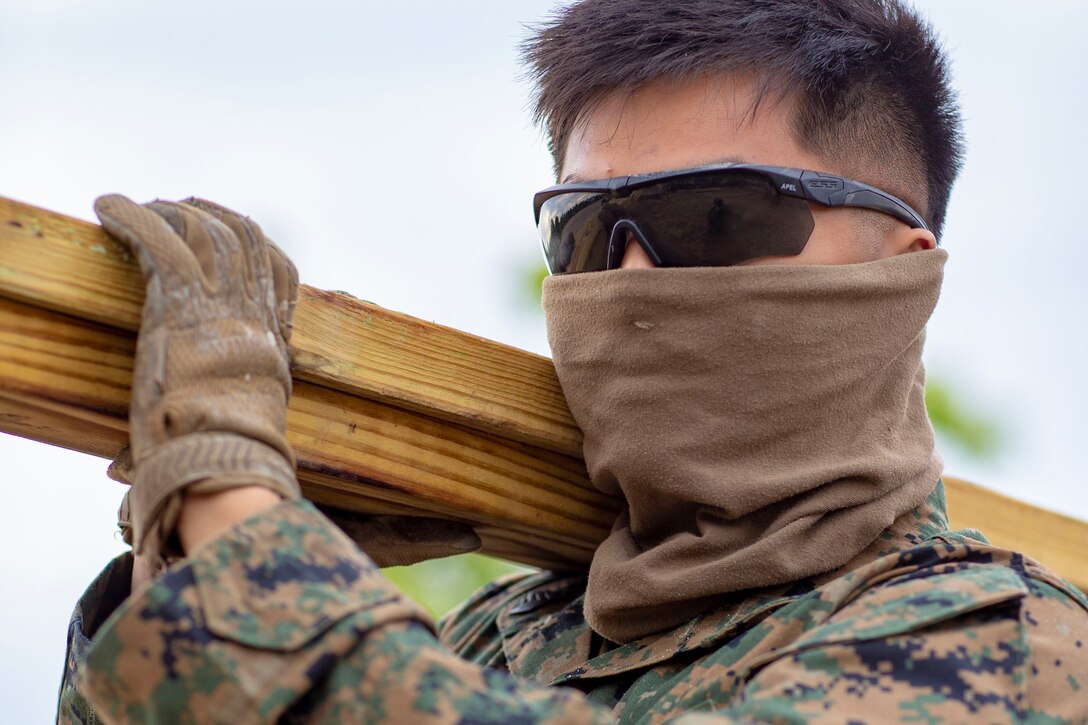 A Marine wearing a face mask carries a piece of lumber over his shoulder.