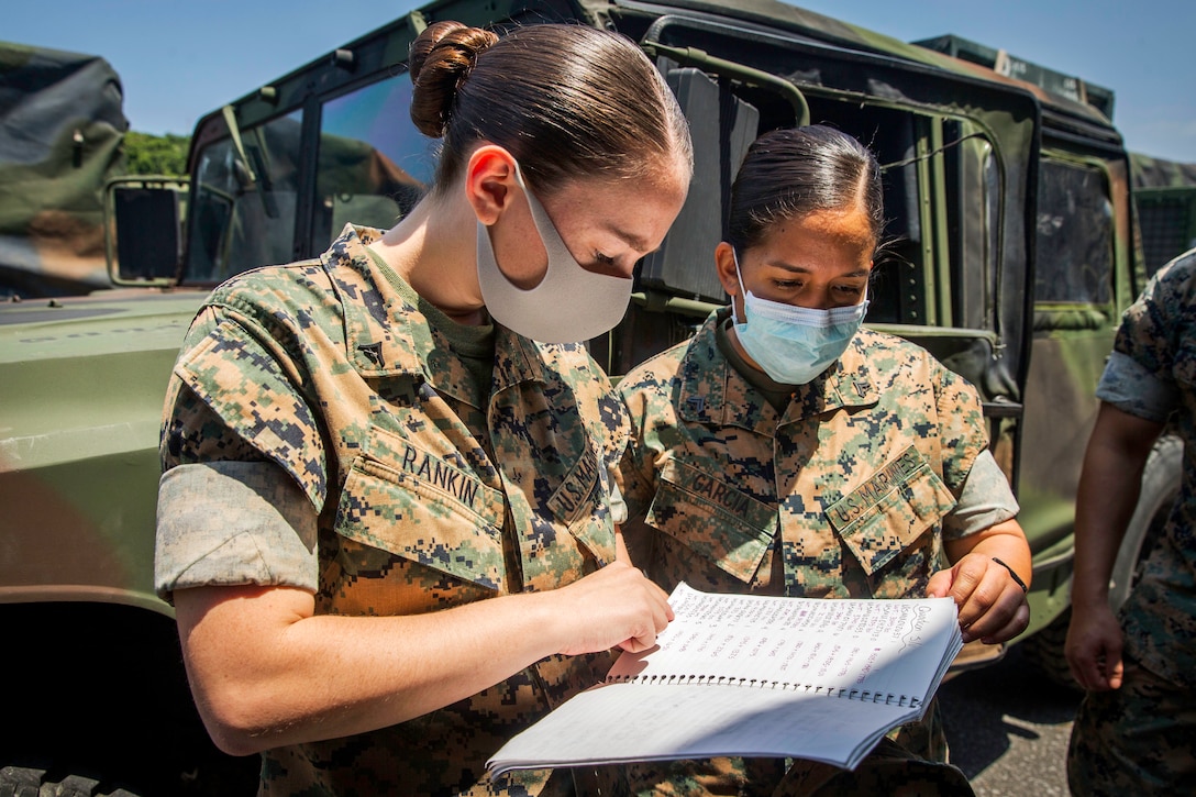 One Marine speaks to another Marine while checking material written in a notebook. Both wear medical face masks. A jeep is in the background.