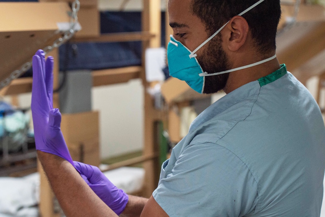 A seaman wearing hospital scrubs and a face mask puts on a pair of gloves.