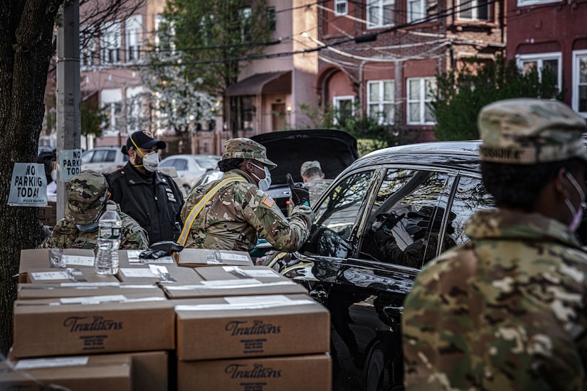 Soldiers give out boxes of food on a crowded urban street.