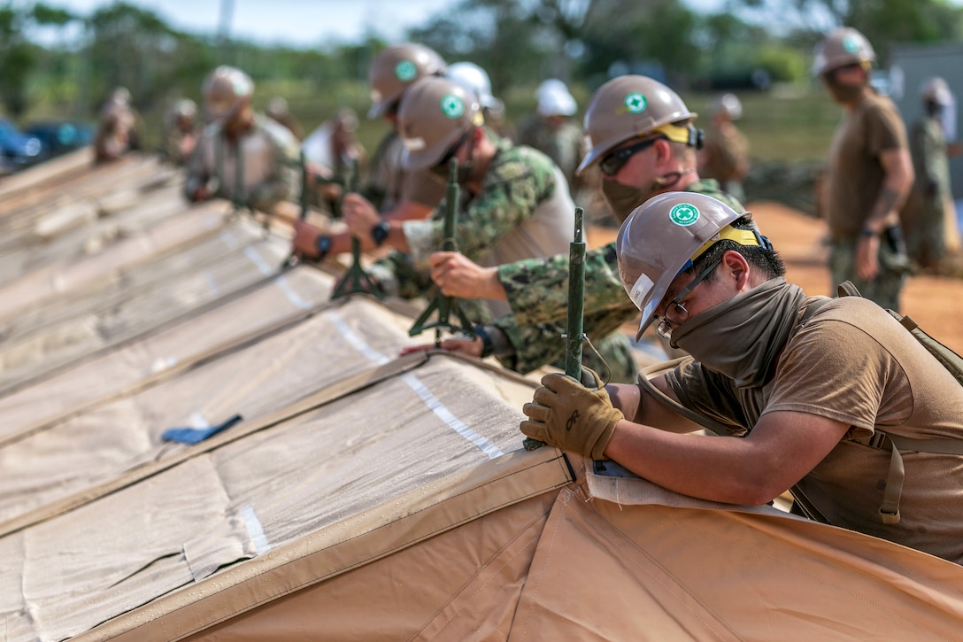 Sailors wearing hard hats and face masks work in a line on a tent.