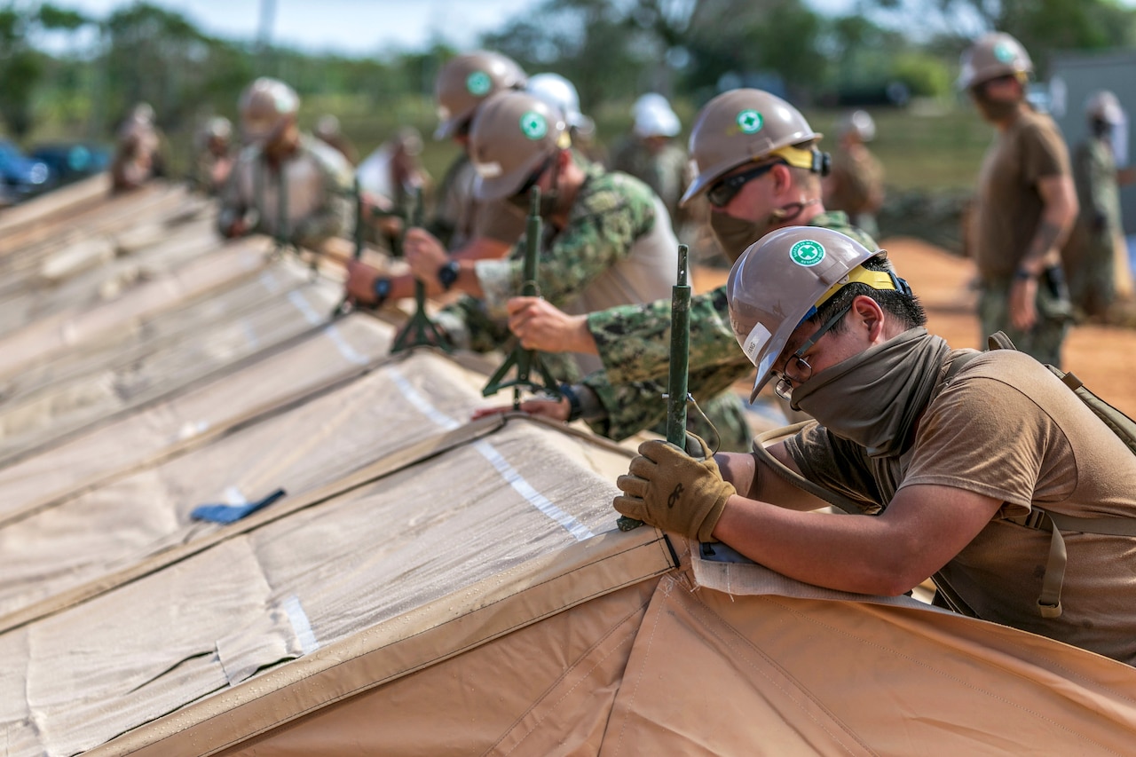 Sailors wearing hard hats and face masks work in a line on a tent.
