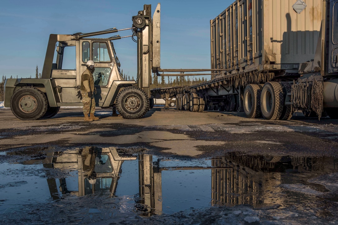 An airman stands by a forklift that faces a container.