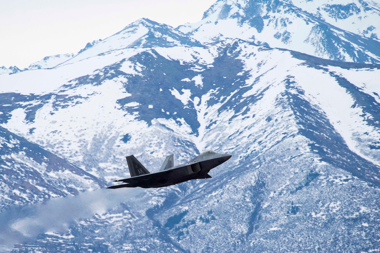 A military aircraft flies in front of snow-covered mountains.