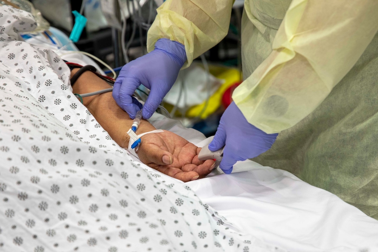 Hands in purple gloves adjust medical equipment on a patient’s arm.