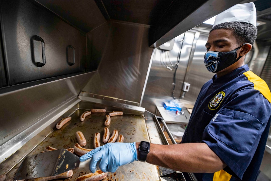 A sailor wearing a face mask and gloves cooks sausages on a griddle.