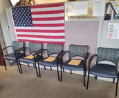 Five blue chairs with yellow envelopes on three chairs are lined up against a pink and white cubicle wall with an American flag.