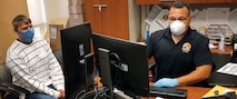 White male in white and black striped shirt wears a blue face mask sits in an office chair while male in black polo shirt with blue gloves and white face mask sits behind a desk.