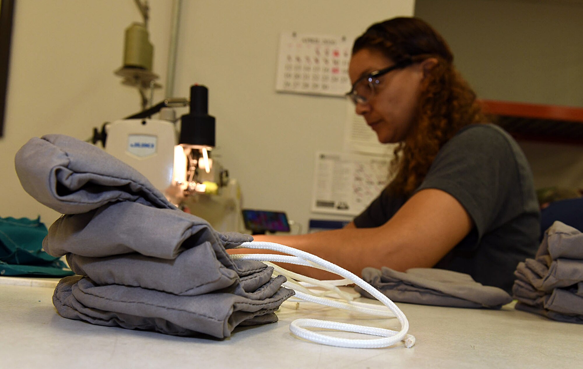 Photo shows a close up of a stack of masks with a woman in the background working a sewing machine.