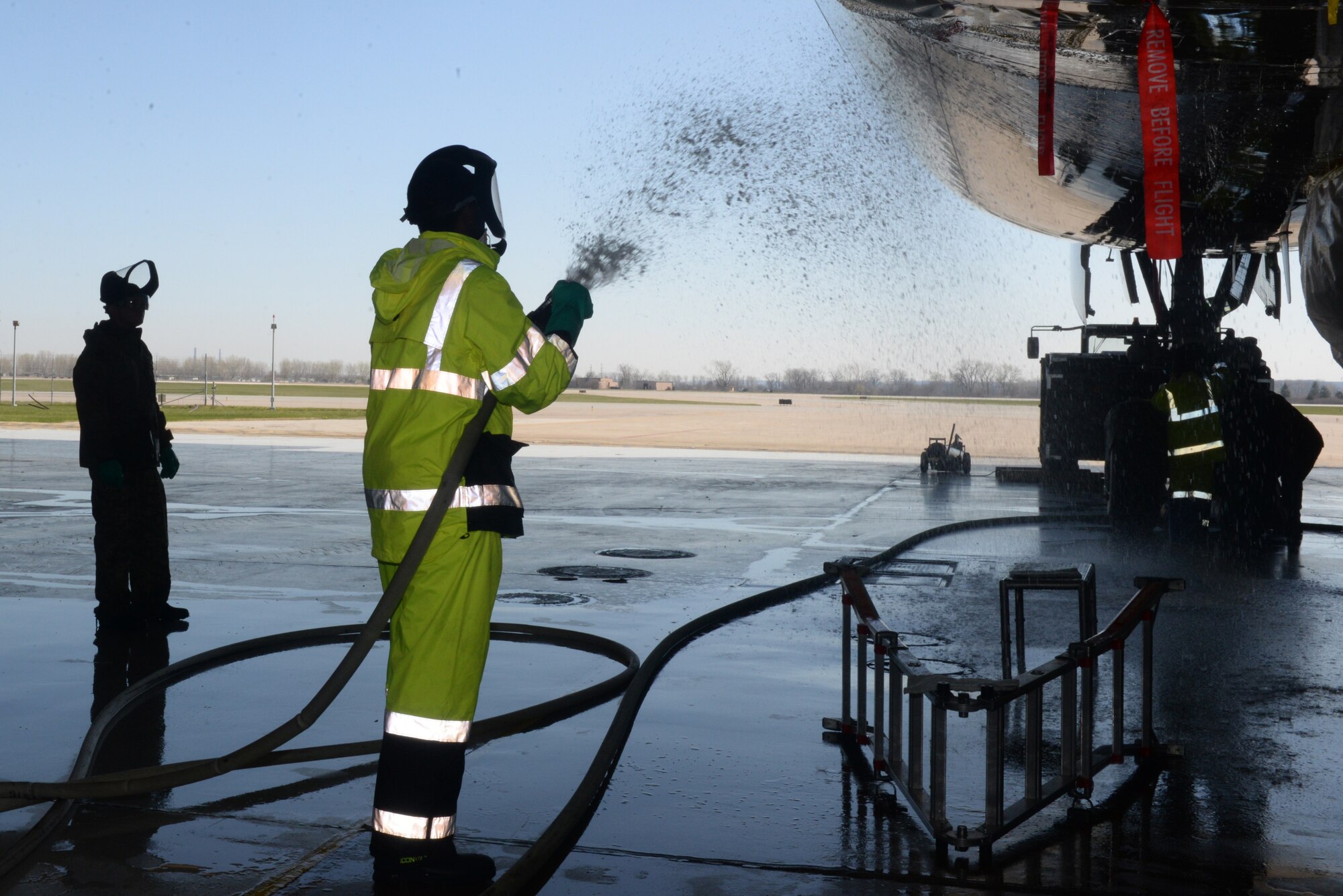 Airmen spray an E4B using a fire hose connected to a fire hydrant.