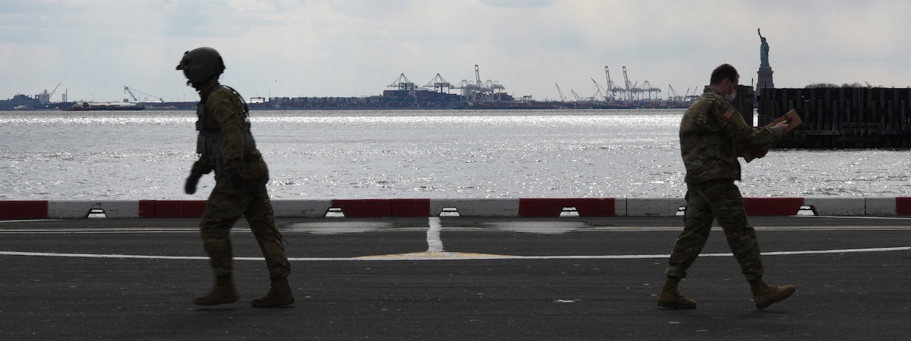 A crew member walks toward the left to return to his helicopter as a soldier walking toward the right carries a package the crew member delivered.