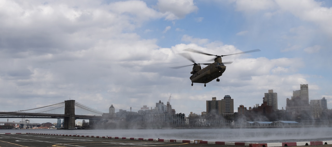 A helicopter with two top rotors nears a landing pad. A bridge and the New York City skyline are in the background.