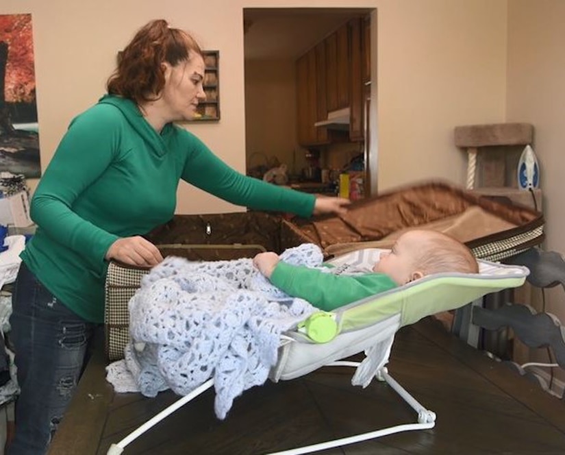 A mother opens a suitcase on a table beside a baby in a baby seat.