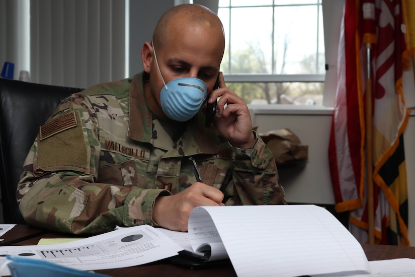 A soldier wearing a face mask writes as he talks on a telephone.