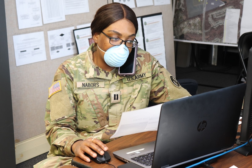 Masked soldier talks on a cellphone at a desk.