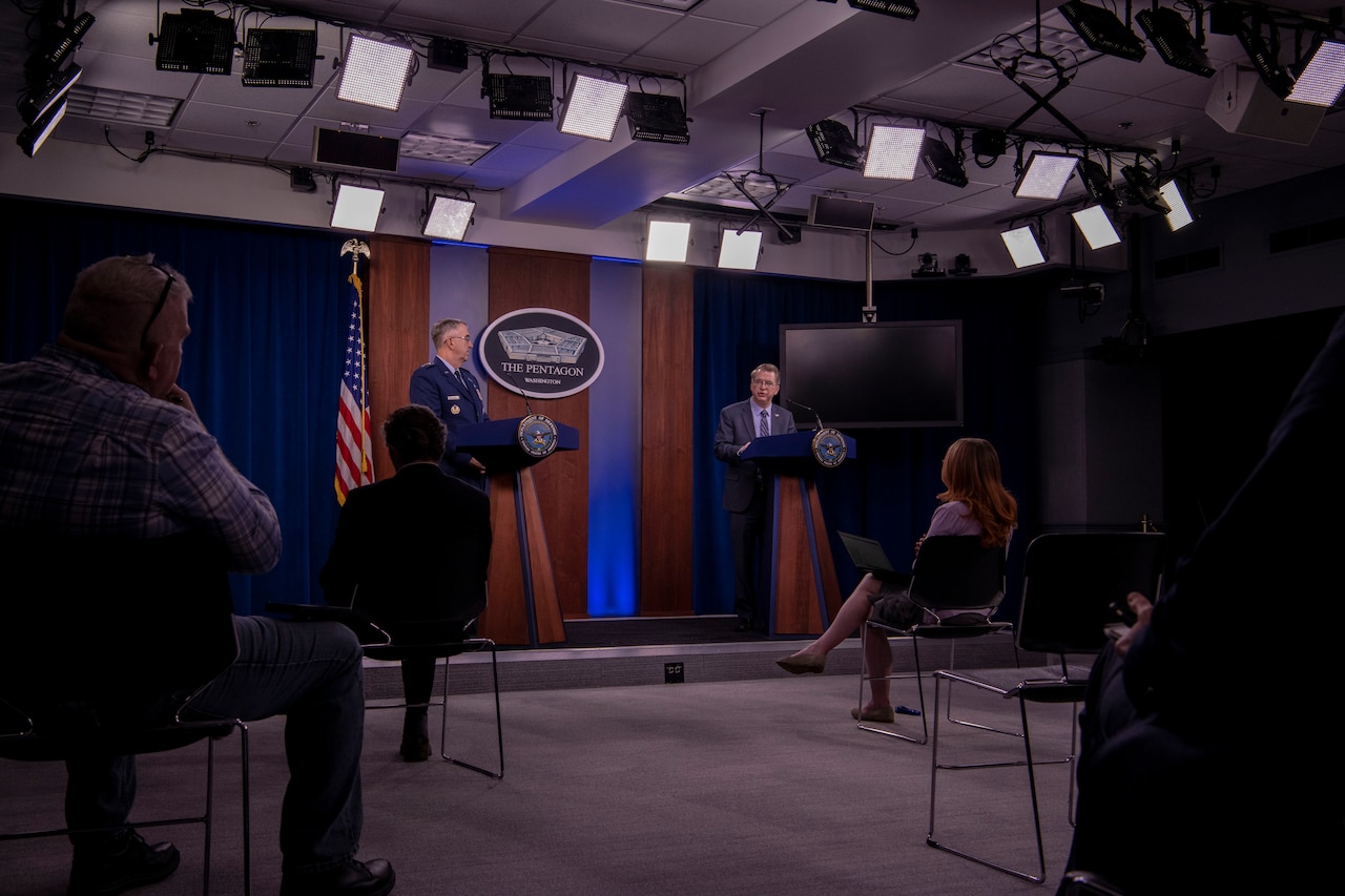 Two men stand behind lecterns and speak to people who are social distancing.