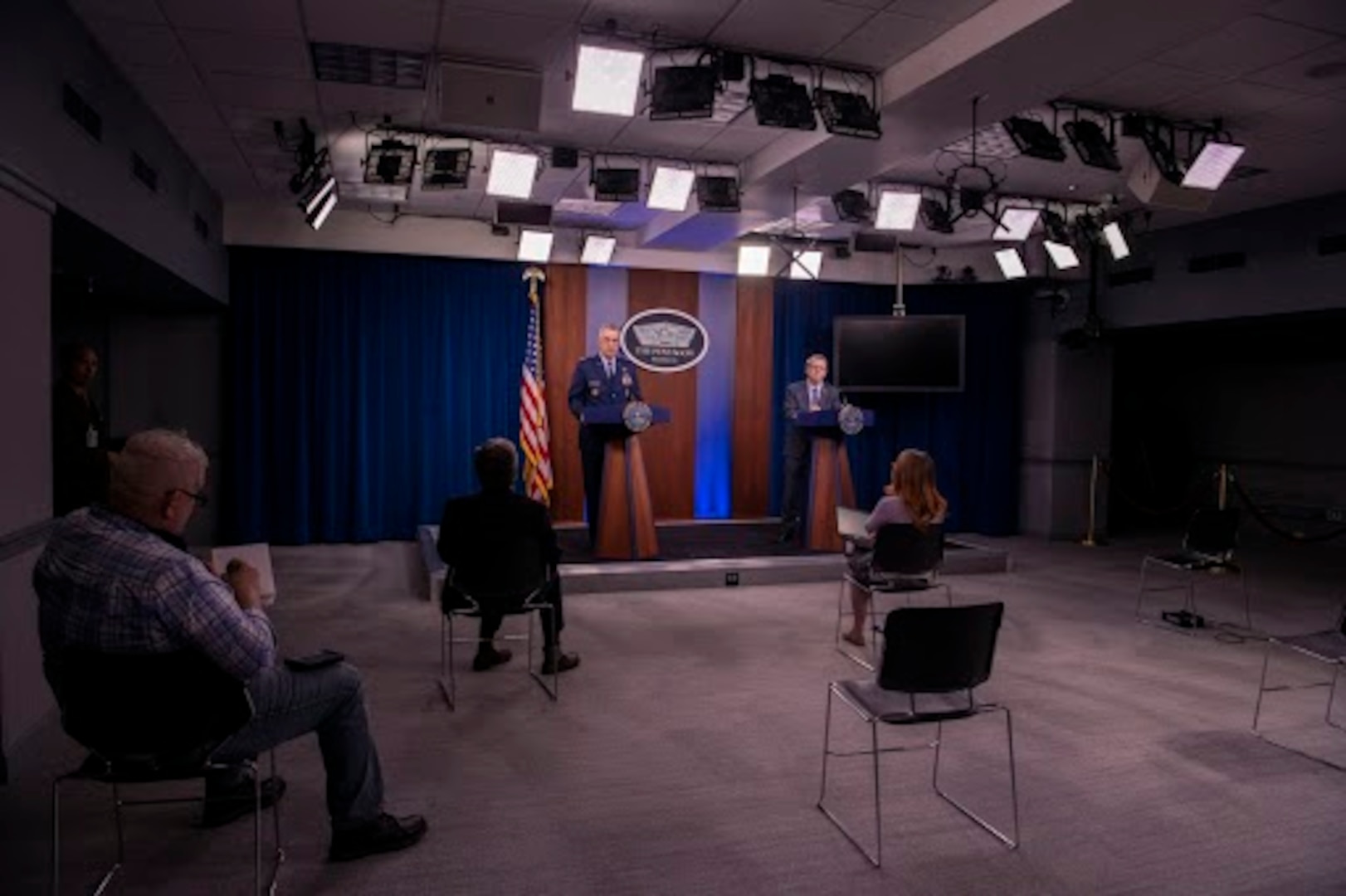 Two men speak from a stage at a news conference, with reporter seating that adheres to social distancing guidelines.