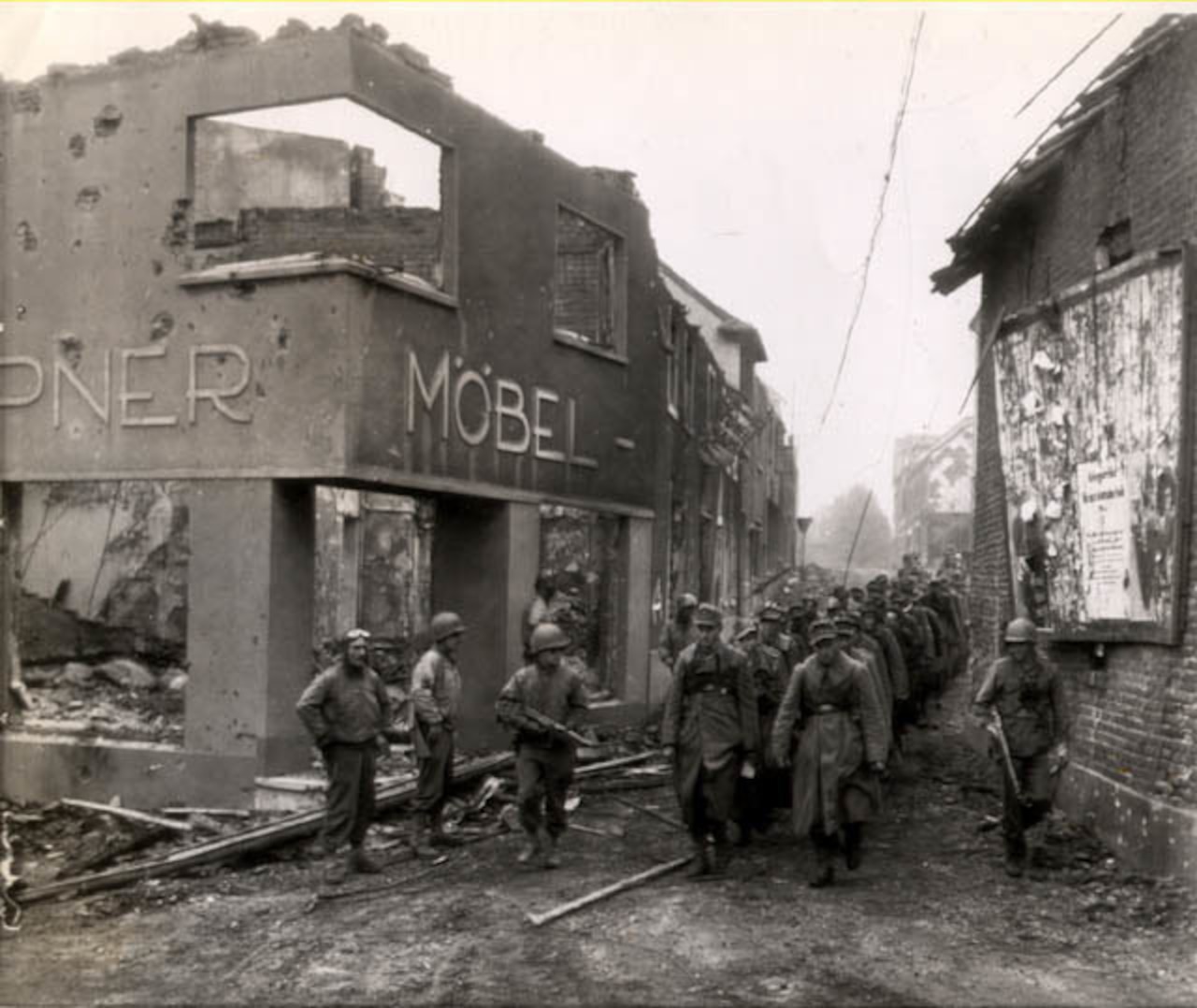 Soldiers guarded by four others march in two lines through a street lined with destroyed buildings.