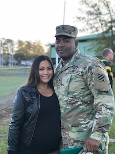 Pacific-American female in black leather jacket and black shirt stands next to black male in green camouflage uniform and cap.