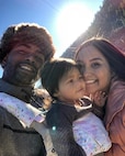Black male in fur hat stands next to young pacific American daughter and wife in pink jacket. Sun is shining in background.