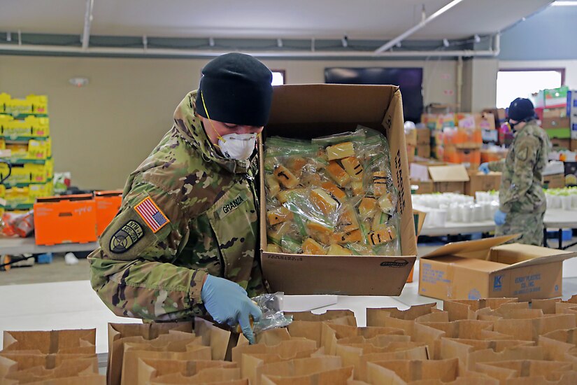 A soldier wearing personal protective equipment adds items from a box he is holding to dozens of small paper bags on a counter.