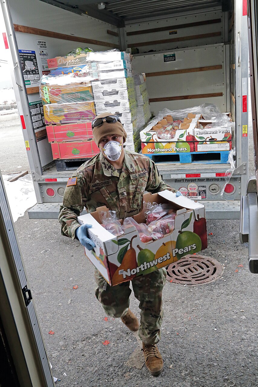 A soldier wearing personal protective equipment carries a box of fruit from a delivery truck.