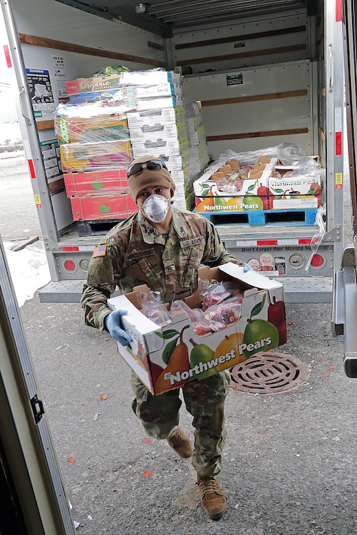 A soldier wearing personal protective equipment carries a box of fruit from a delivery truck.