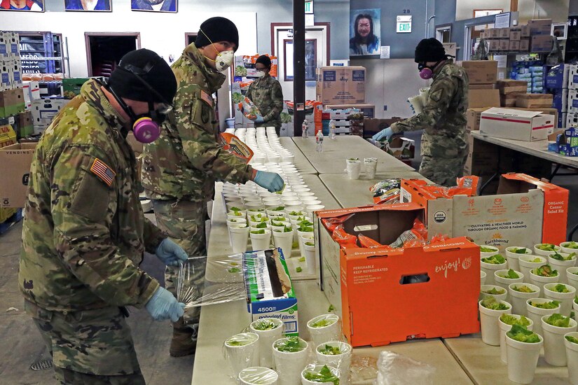Soldiers wearing personal protective equipment assemble packaged meals.