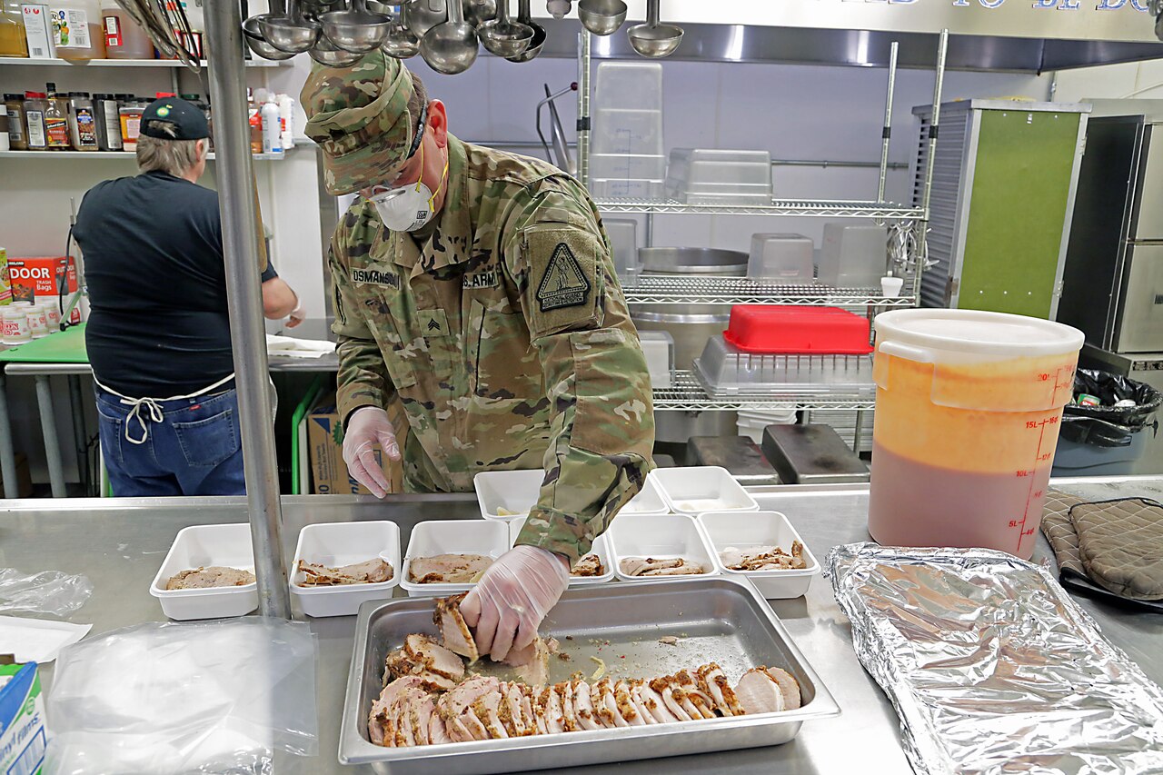 A soldier wearing personal protective equipment loads sliced turkey into food containers.
