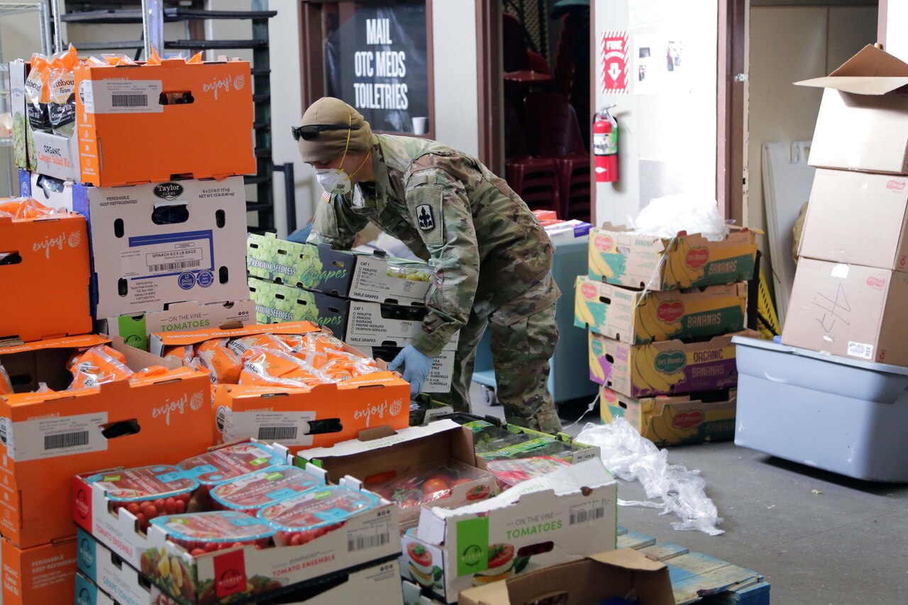 A soldier wearing personal protective equipment stacks boxes of food.