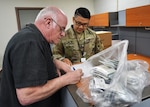 A man signs a piece of paper with a bag of unrecognizable objects next to him as a service member in uniform watches.
