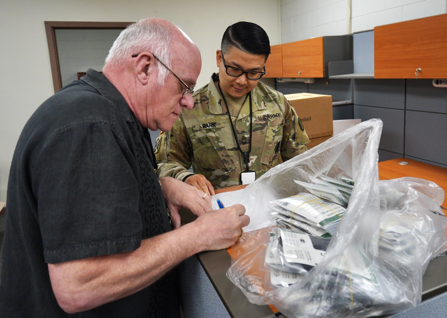 A man signs a piece of paper with a bag of unrecognizable objects next to him as a service member in uniform watches.