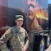 White male in green camouflage uniform in cap stands next to a semi trailer with the picture of a Soldier on it.