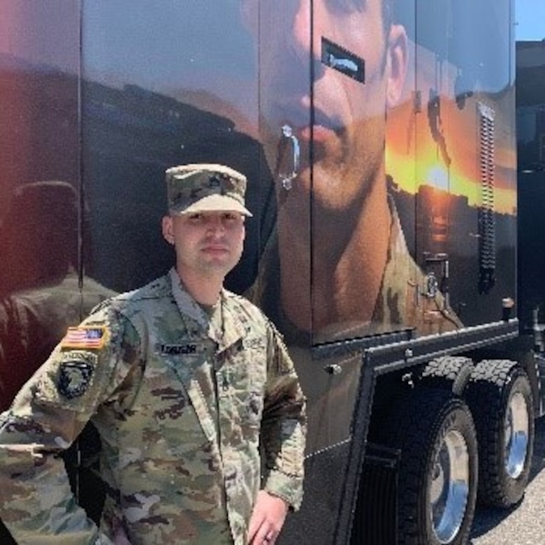 White male in green camouflage uniform in cap stands next to a semi trailer with the picture of a Soldier on it.